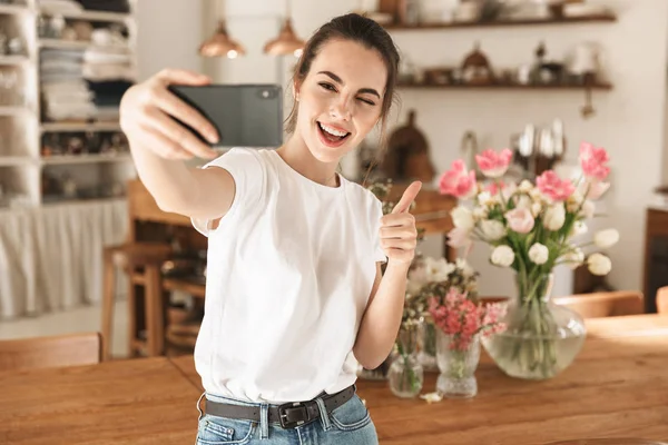 Beyaz tişörtlü güzel genç öğrenci kız kapalı mekanlarda poz vererek tamam jesti göstererek cep telefonuyla selfie çekti.