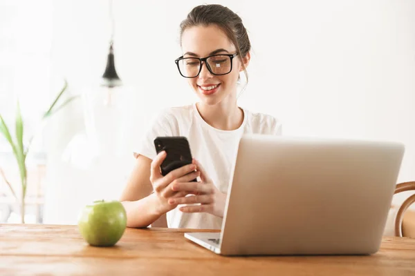 Schöne junge Studentin sitzt drinnen mit Laptop und Handy. — Stockfoto