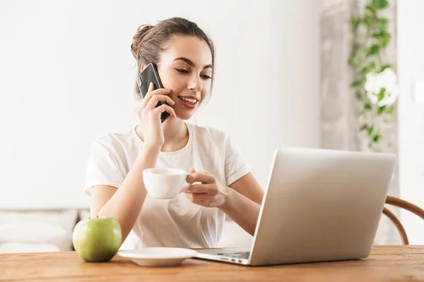 Studente ragazza con mela seduta al chiuso utilizzando computer portatile parlando con il telefono cellulare . — Foto Stock