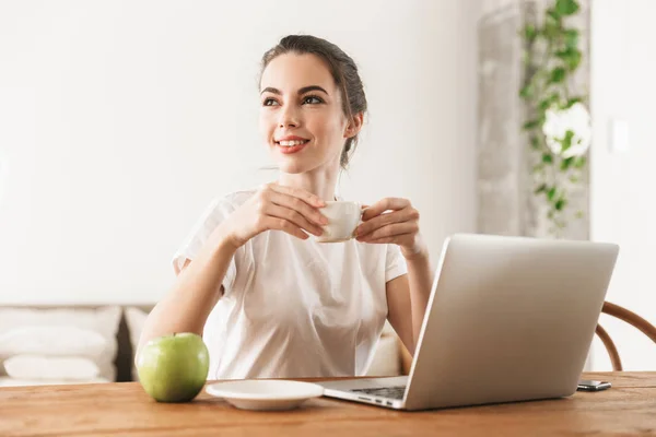 Vacker ung student tjej med äpple sittande inomhus med bärbar dator dricka kaffe. — Stockfoto