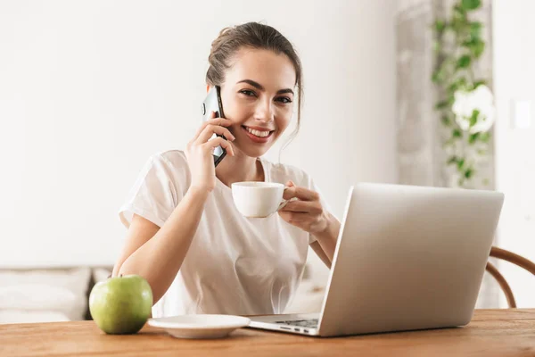 Studentin mit Apfel sitzt drinnen mit Laptop-Computer und telefoniert mit Handy. — Stockfoto