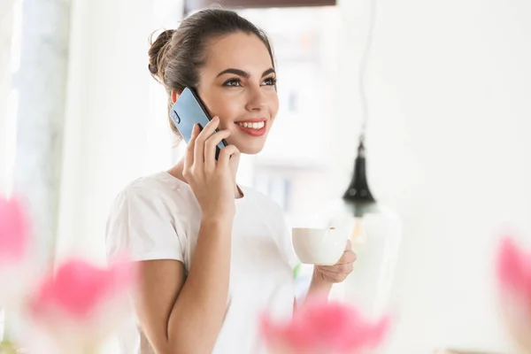 Beautiful young student girl drinking coffee posing indoors talking by mobile phone. — Stock Photo, Image