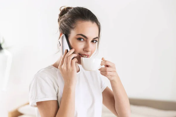 Student meisje drinken koffie poseren binnenshuis praten via mobiele telefoon. — Stockfoto