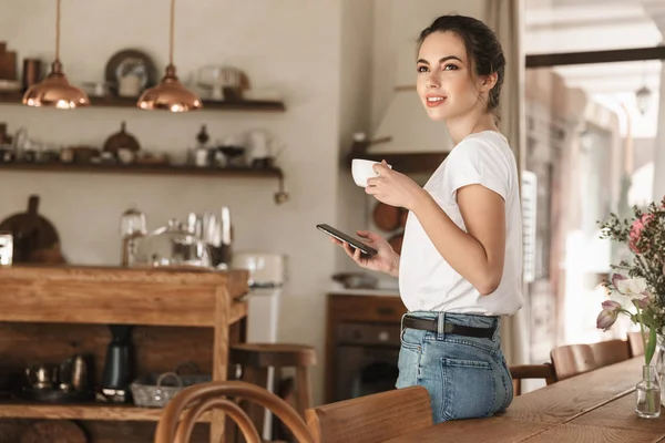 Mooie jonge student meisje drinken koffie poseren binnenshuis chatten via mobiele telefoon. — Stockfoto