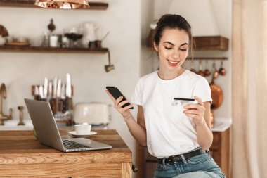 Genç bir öğrenci evde oturup dizüstü bilgisayarla cep telefonuyla kredi kartı tutarak sohbet ediyor..