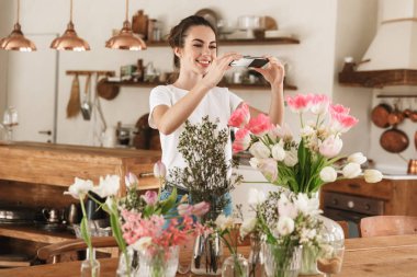 Çiçeklerin yanında poz veren mutlu genç öğrenci kız cep telefonuyla fotoğraf çekmek.