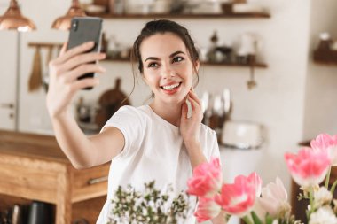 Çiçeklerin yanında poz veren mutlu genç öğrenci kız cep telefonuyla fotoğraf çekmek.