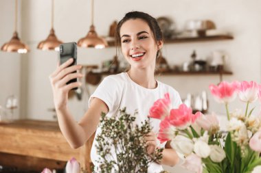 Çiçeklerin yanında poz veren mutlu genç öğrenci kız cep telefonuyla fotoğraf çekmek.