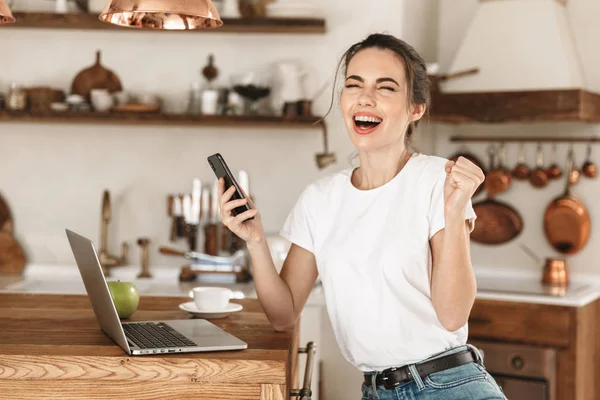 Schöne junge Studentin sitzt drinnen mit Laptop-Computer im Chat per Mobiltelefon. — Stockfoto