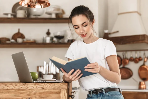 Ung student flicka sitter inomhus med hjälp av bärbar dator läsa bok. — Stockfoto