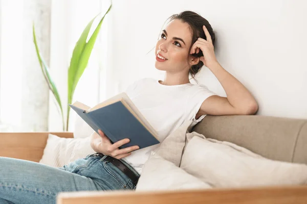 Schöne junge Studentin sitzt drinnen und liest Buch auf dem Sofa. — Stockfoto