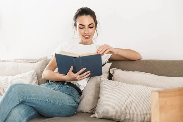 Schöne glückliche junge Studentin sitzt drinnen und liest Buch auf dem Sofa. — Stockfoto