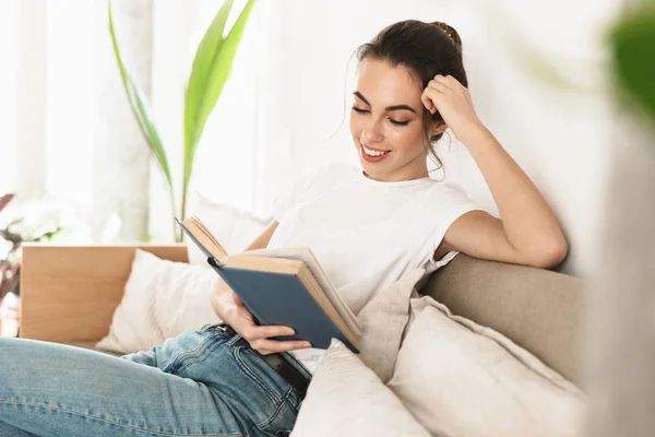 Piękny szczęśliwy młody student dziewczyna siedzący w pomieszczeniu czytanie książka na sofa. — Zdjęcie stockowe