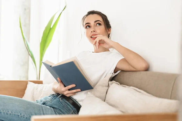 Schöne junge Studentin sitzt drinnen und liest Buch auf dem Sofa. — Stockfoto