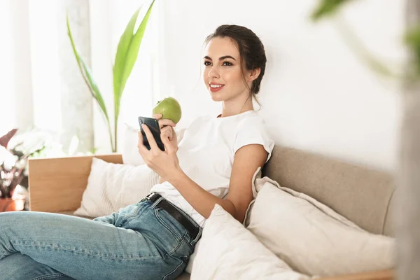 Happy young student girl sitting indoors eat apple using mobile phone on sofa. — Stock Photo, Image