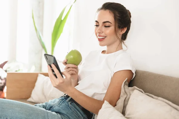 Счастливая студентка сидит в помещении и ест яблоко с мобильного телефона на диване. . — стоковое фото