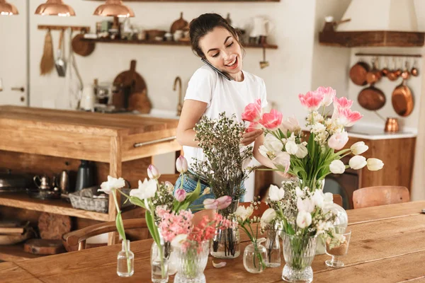 Beautiful happy young student girl posing indoors near flowers talking by mobile phone. — Stock Photo, Image