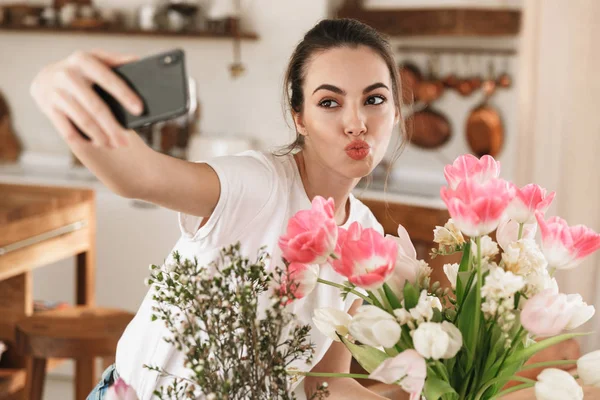 Çiçeklerin yanında poz veren mutlu genç öğrenci kız cep telefonuyla fotoğraf çekmek. — Stok fotoğraf