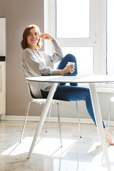 Smiling young woman drinking coffee while sitting at the table — Stock Photo, Image