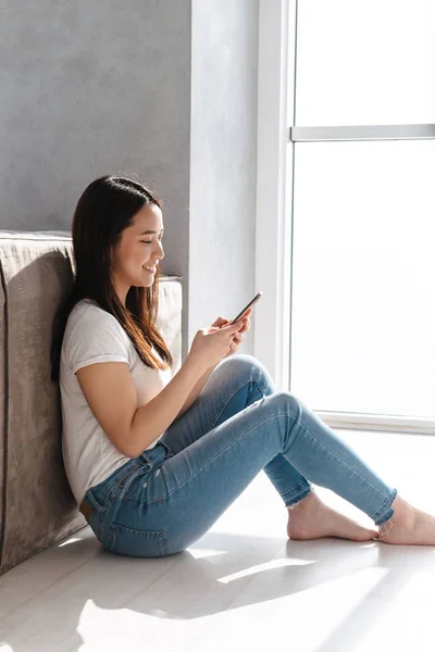 Portrait of satisfied asian girl using smartphone while sitting — Stock Photo, Image