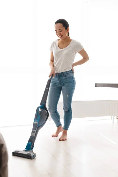 Retrato de alegre asiático mulher 20s fazendo tarefas domésticas com vácuo c — Fotografia de Stock