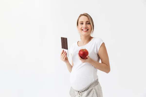 Mooie jonge zwangere vrouw poseren geïsoleerd over witte muur achtergrond houden van appel en chocolade. — Stockfoto
