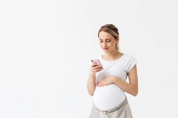 Feliz joven embarazada posando aislado sobre fondo blanco de la pared utilizando el teléfono móvil . — Foto de Stock