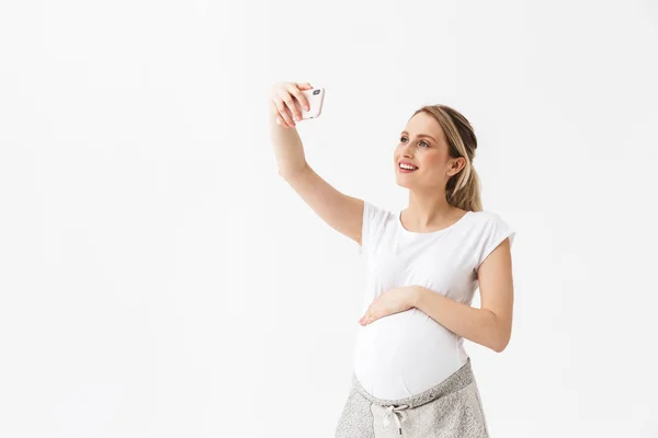 Щаслива молода вагітна жінка позує ізольовано на білому тлі стіни візьміть селфі на мобільний телефон . — стокове фото