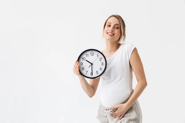 Feliz joven embarazada posando aislado sobre fondo blanco de la pared celebración del reloj . —  Fotos de Stock