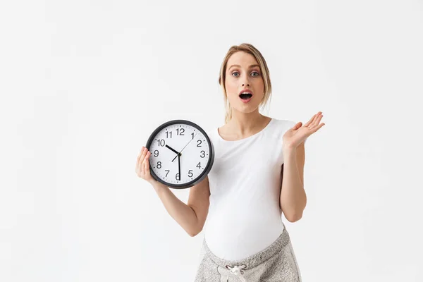 Шокированная молодая беременная женщина позирует на белом фоне, держа часы . — стоковое фото