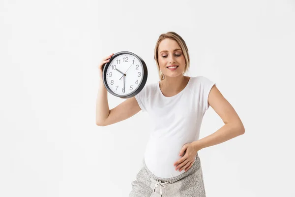 Feliz joven embarazada posando aislado sobre fondo blanco de la pared celebración del reloj . —  Fotos de Stock