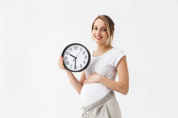 Gelukkige jonge zwangere vrouw poseren geïsoleerd over witte muur achtergrond Holding klok. — Stockfoto