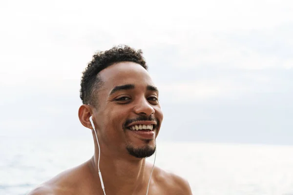 Feliz bonito jovem homem de esportes africanos ao ar livre na praia mar andando ouvindo música com fones de ouvido . — Fotografia de Stock