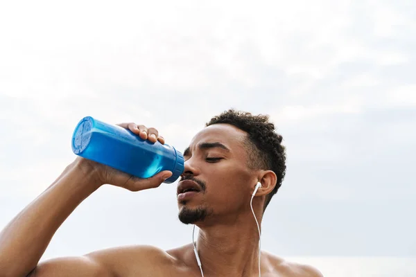 強いハンサムな若いアフリカのスポーツマンは、イヤホンで音楽を聴いてビーチ海で水を飲みます. — ストック写真