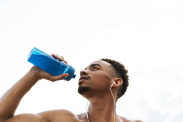 強いハンサムな若いアフリカのスポーツマンは、イヤホンで音楽を聴いてビーチ海で水を飲みます. — ストック写真