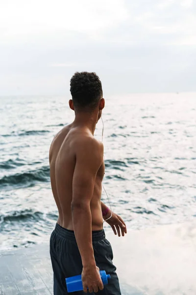 Yakışıklı genç Afrikalı spor adam plaj denizde açık havada su ile şişe tutan kulaklık ile müzik dinliyor. — Stok fotoğraf