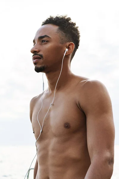 Bonito jovem homem de esportes africanos ao ar livre na praia mar ouvir música com fones de ouvido . — Fotografia de Stock