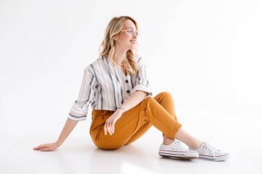 Bir kenara bakan ve yerde oturan gözlük takan sarışın gülümseyen kadın portresi