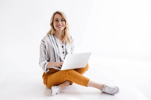 Imagem de uma jovem mulher usando óculos sorrindo para a câmera e usando laptop enquanto sentada no chão — Fotografia de Stock