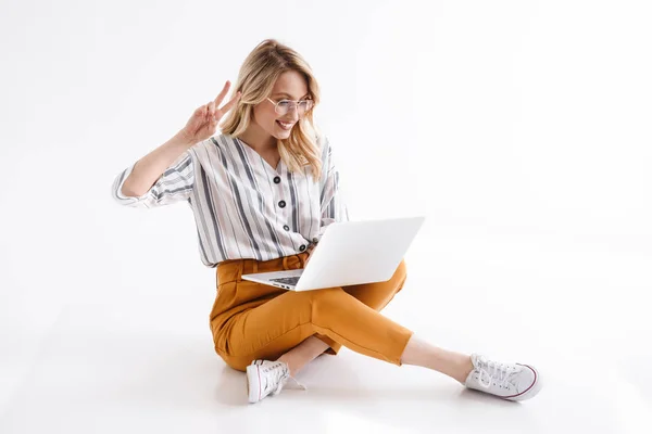 Immagine di una donna bionda che indossa occhiali usando il computer portatile e mostra la pace cantare mentre è seduta sul pavimento — Foto Stock