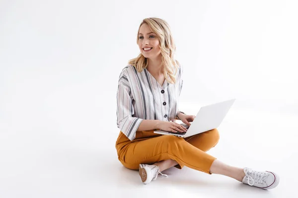 Immagine di una bella donna sorridente che indossa abiti casual digitando sul computer portatile e guardando da parte mentre si siede sul pavimento — Foto Stock