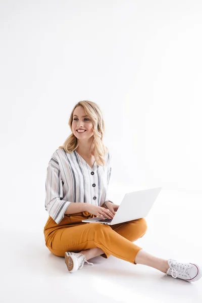 Imagen de una linda mujer sonriente usando ropa casual escribiendo en el portátil y mirando a un lado mientras está sentada en el suelo — Foto de Stock