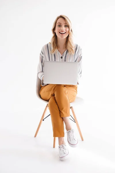 Foto de mujer linda caucásica usando ropa casual sonriendo a la cámara y usando el ordenador portátil mientras está sentado en la silla — Foto de Stock