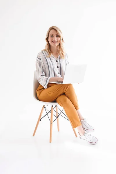 Foto de una buena mujer europea que usa ropa casual sonriendo a la cámara y usando un portátil mientras está sentada en la silla — Foto de Stock