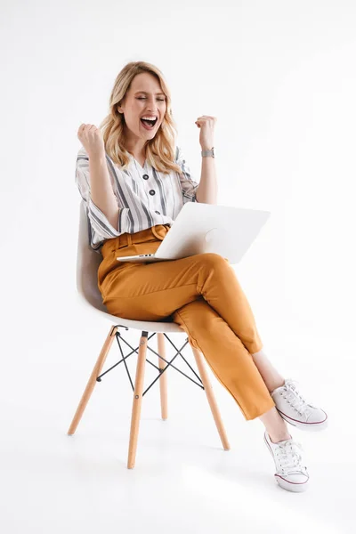 Foto di una donna felice europea che indossa abiti casual usando il computer portatile mentre è seduta sulla sedia — Foto Stock