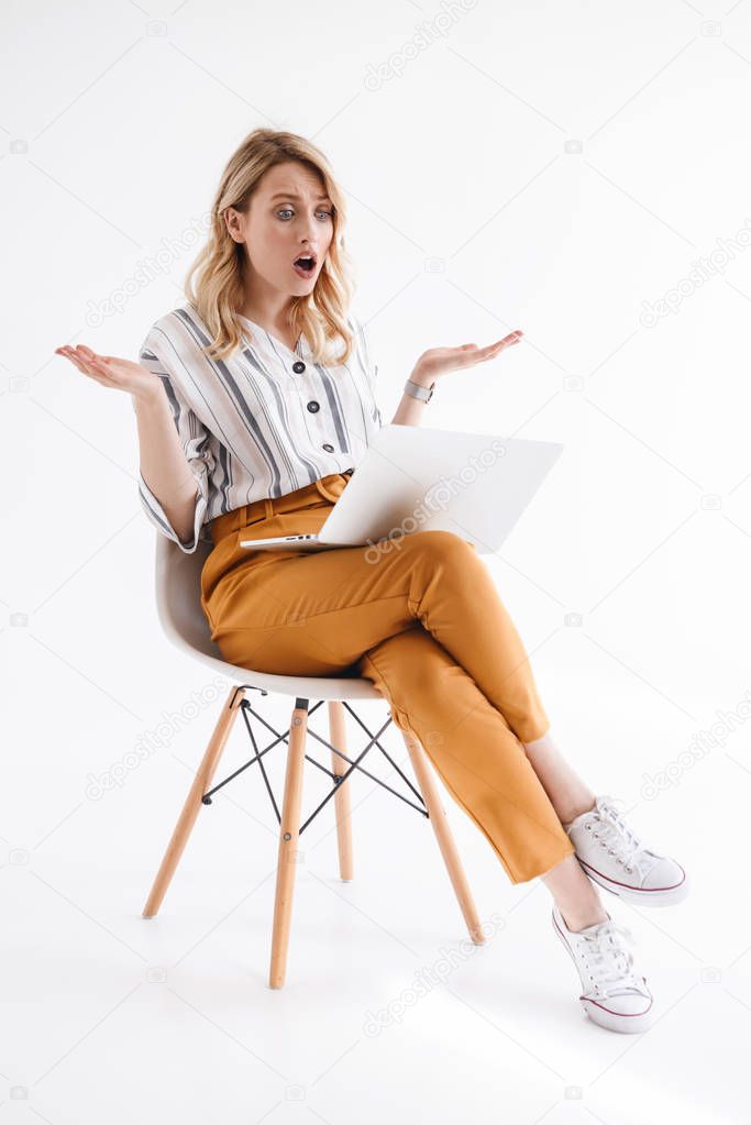 Portrait of shocked girl wearing casual clothes looking at laptop and throwing up hands while sitting in chair