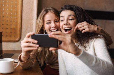 Heyecanlı mutlu güzel kız arkadaşlar cep telefonu kullanarak kafede oturan.