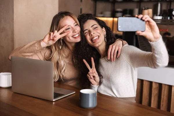 Dizüstü bilgisayar kullanarak kafede oturan heyecanlı mutlu kız arkadaşlar telefonla selfie çekmek. — Stok fotoğraf