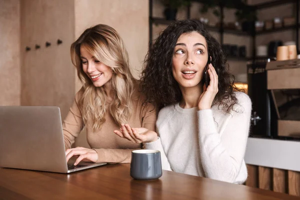 Возбужденные эмоциональные девушки друзья сидят в кафе, используя ноутбук, разговаривая по мобильному телефону . — стоковое фото