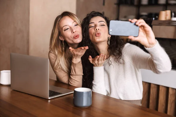 Dizüstü bilgisayar kullanarak kafede oturan heyecanlı mutlu kız arkadaşlar telefonla selfie çekmek. — Stok fotoğraf